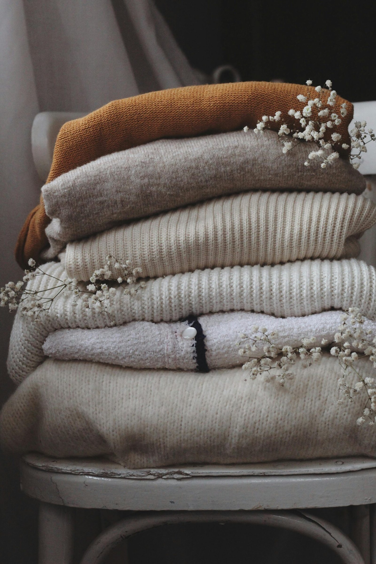 Aesthetic stack of beige and brown woolen sweaters on a chair indoors, adorned with dried flowers.